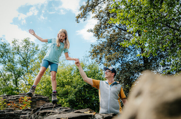 Das Bild fängt einen Moment der Freude und des Abenteuers zwischen einem Mann und einem jungen Mädchen ein. Sie stehen auf einer felsigen Erhebung, wobei das Mädchen spielerisch von der Kante springt. Der Mann, der eine gelbe Weste trägt, hält ihre Hand, um ihre Sicherheit zu gewährleisten. Das Mädchen, das ein blaues Hemd und Shorts trägt, schwebt in der Luft, ihre Arme ausgebreitet, als ob sie flöge. Der Hintergrund ist ein üppiger grüner Wald, mit Bäumen und Büschen, die das Landschaftsbild zieren. Der Himmel über ihnen ist ein klares Blau, das zur Gesamtstimmung des Bildes beiträgt. Das Bild strahlt Freiheit, Erkundung und die einfachen Freuden der Kindheit aus.