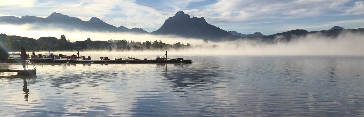 Das Bild fängt eine friedliche Szene eines kleinen grünen Bootes ein, das sanft auf dem Hopfensee schwimmt. Das Boot befindet sich im Vordergrund, hinter ihm breitet sich der weite Horizont des Wassers aus. Im Hintergrund erhebt sich die majestätische Bergkette der Ammergauer Alpen, die teilweise von einer Schicht Nebel verdeckt sind. Der Himmel über ist ein klares Blau, durchsetzt mit einigen Wolken. 