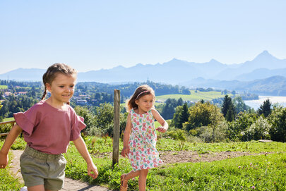 Das Bild fängt einen friedlichen Moment in einem Park ein, wo zwei junge Mädchen einen Spaziergang genießen. Das Mädchen auf der linken Seite, in einem pinkfarbenen Shirt und olivgrünen Shorts gekleidet, marschiert voraus, während das zweite Mädchen, das ein Blumenkleid trägt, dicht hinter ihr herläuft. Sie laufen auf einem Pfad, der von üppigem Grün und Bäumen gesäumt ist. Als Hintergrund des Bildes dient eine atemberaubende Aussicht auf einen See und Berge, was zur Ruhe der Szene beiträgt. Die Mädchen scheinen guter Stimmung zu sein, denn sie beide lächeln. Die Gesamtatmosphäre des Bildes ist friedlich und fröhlich.