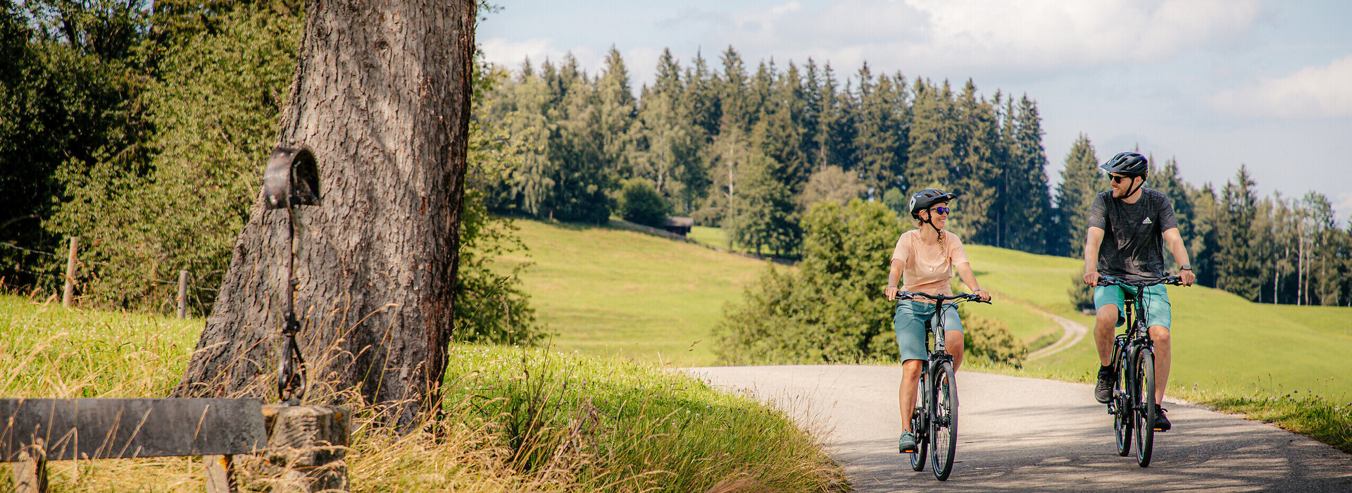 Das Bild zeigt einen Mann und eine Frau beim Fahrradfahren. Beide tragen Freizeitkleidung und einen Helm. Sie fahren auf den Betrachter zu. Sie lächeln einander an. Beide tragen Sonnenbrillen. Die Straße, auf der sie fahren ist asphaltiert. Wiesen und Wälder säumen die Straße. Am Straßenrand ist eine Wegkreuz aus Metall und eine Holzbank zu sehen.  Im Hintergrund erheben bewaldete Hügel. Der Himmel ist blau. Die Sonne scheint. 