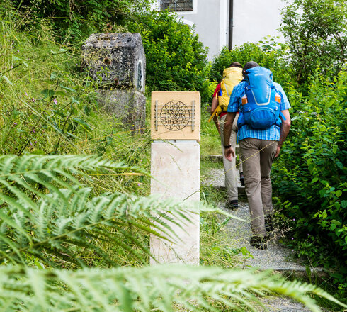 Das Bild zeigt einen Mann und eine Frau in Wanderkleidung mit Rucksäcken. Sie wandern einen Hang empor auf einem Kiesweg mit Stufen. Im Vordergrund steht eine steinerne Säule. Um sie herum wachsen Büsche, Bäume und Gräser.   