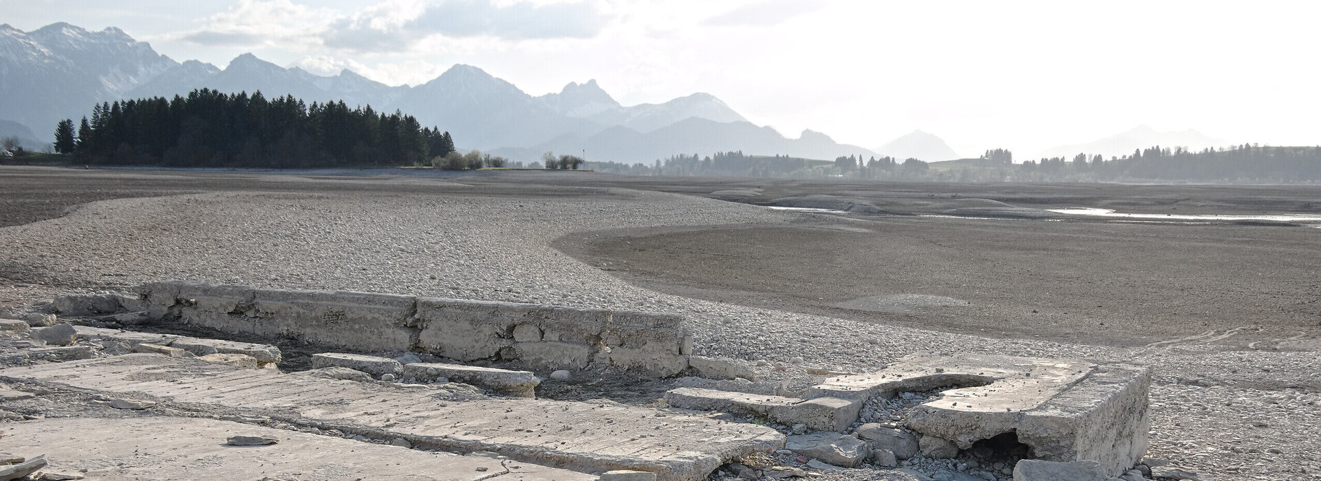 Das Foto zeigt das leere Bett des Forggensees. Nur Steine und Kies sind zu sehen. Im Hintergrund erheben sich die Alpen. Davor stehen grüne Wälder. Im Vordergrund ist ein altes, zerstörtes Betonfundament zu sehen.
