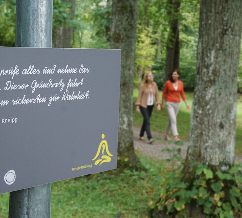 Das Bild zeigt zwei Frauen, die auf einem Waldweg laufen. Ringsherum wachsen hohe Bäume. Auf dem Schild im Vordergrund steht „Man prüfe alles und nehme das Beste. Dieser Grundsatz führt uns am sichersten zur Wahrheit. Sebastian Kneipp“