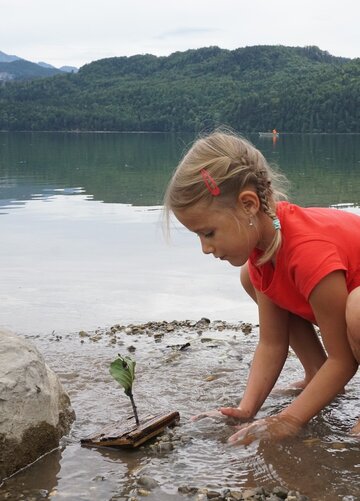 Das Bild zeigt eine Mädchen mit zwei Zöpfen, dass am Ufer eines Sees spielt. Sie ist in die Hocke gegangen, barfuß, trägt kurze Freizeitkleidung und hat die Hände im Wasser. Im Hintergrund erheben sich bewaldete Hügel und hohe Berge. 