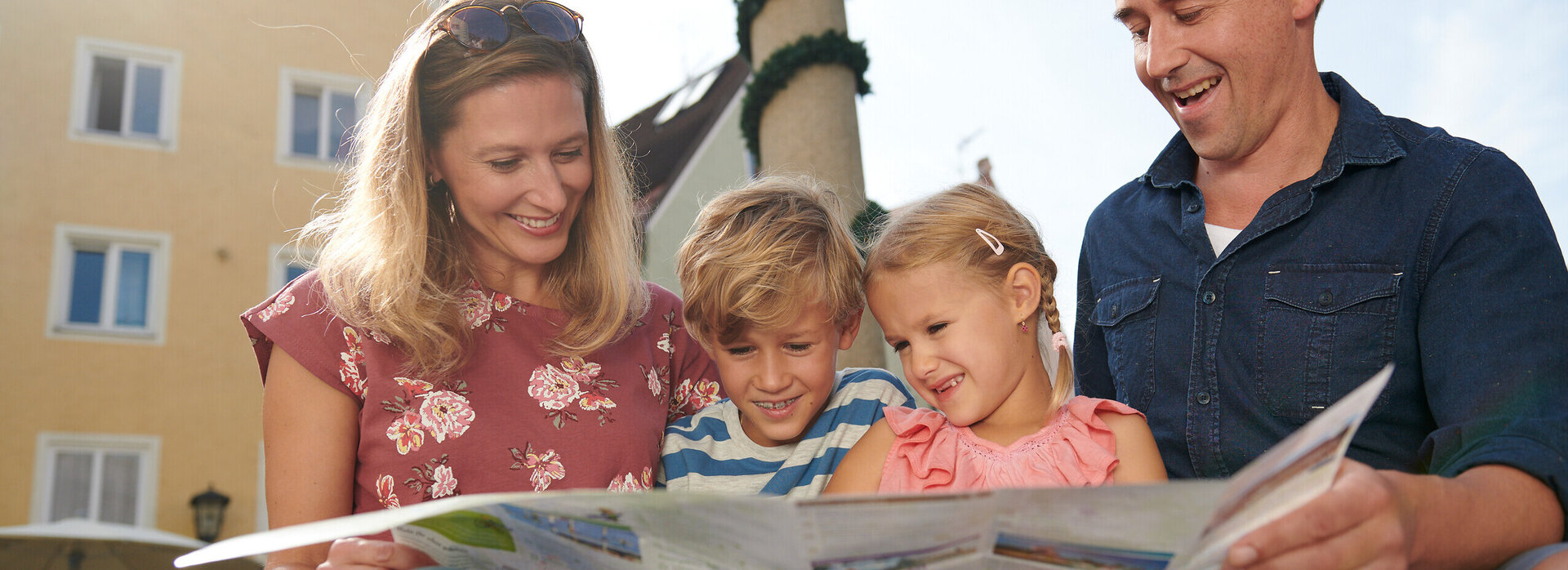  Das Bild ist ein Farbfoto, das eine Familie von vier Personen zeigt, die draußen sitzen. Die Frau, die wahrscheinlich die Mutter ist, ist auf der linken Seite des Bildes zu sehen, sie sitzt und hält eine Zeitung aufgeschlagen vor sich. Sie lächelt und sieht in die Kamera. Neben ihr sitzt ein junges Mädchen, ebenfalls lächelnd und in die Kamera blickend. Das Mädchen trägt ein Rosa-Top. Auf der rechten Seite des Bildes sitzt ein Mann, der ebenfalls die Zeitung aufgeschlagen vor sich hält. Er lächelt und sieht in die Kamera. Der Mann trägt ein Blau-Shirt. Im Hintergrund ist ein gelbes Gebäude mit mehreren Fenstern zu sehen. Der Himmel ist klar und blau, was auf einen sonnigen Tag hindeutet. Es sind keine sichtbaren Texte oder Logos im Bild zu sehen.
