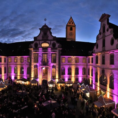 Das Bild zeigt den Füssener Adventsmarkt im Klosterhof des ehemaligen Benediktinerklosters St. Mang. Die Fassade des rundumlaufenden Gebäudes ist pink beleuchtet. Die Buden des Adventsmarkt sind ebenfalls festlich beleuchtet. Zahlreiche Menschen sind zu sehen. Gerade ist die Sonne untergegangen. Man sieht noch die letzten Sonnenstrahlen über dem Klosterdach.  
