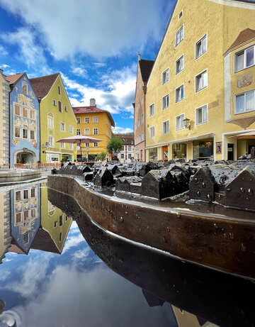 Das Bild zeigt eine malerische Szene eines mit Wasser gefüllten Stadtmodels aus Bronze mit Gebäuden, die sich im Wasser spiegeln. Die Gebäude sind bunt und zeigen die mittelalterliche Stadt von Füssen