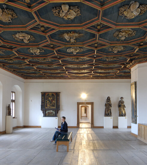 Man blickt in einen Saal mit weißen Wänden, einem historischen Holzboden und einer reich verzierten, dunkelblauen Holzdecke. Das Muster der Decke ist wabenförmig. Die Kanten der Waben sind erhaben und mit roten Umrandungen versehen. Innerhalb jeder Wabe ist eine, in Gold gearbeitete, kirchliche Person zu sehen. Man sieht Heiligenfiguren, aber auch Bischöfe und Päpste. An den Wänden hängen historische Gemälde mit unterschiedlichen Themen. Der Raum ist, bis auf eine moderne mit schwarzem Leder bezogene Bank in der Mitte, nicht möbliert. Auf der Bank sitzt eine Frau und blickt auf die Gemälde. 