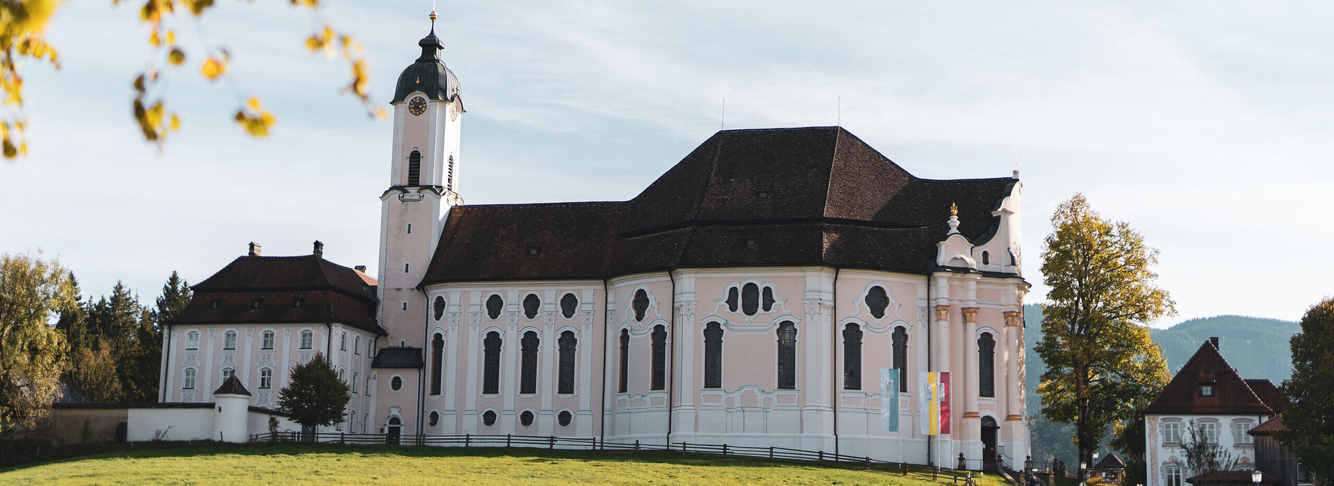 Das Bild zeigt eine friedliche Szene einer großen weißen Kirche mit einem rosa Dach, die majestätisch in der Mitte eines üppigen grünen Feldes steht. Die Kirche, mit ihrem hohen Turm und den vielen Fenstern, strahlt eine Atmosphäre der Ruhe und Ehrfurcht aus. Das Feld, lebendig und grün, erstreckt sich um die Kirche und schafft ein malerisches Setting. In der Ferne sind ein paar Bäume zu sehen, die zur natürlichen Schönheit der Szene beitragen. Die Kirche, mit ihrem auffälligen rosa Dach, hebt sich gegen das grüne Feld ab und zieht die Aufmerksam auf ihre architektonische Pracht. Die Gesamtansicht des Bildes suggeriert eine friedliche und idyllische Szene, vielleicht ein Ort der Anbetung und Reflexion.