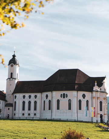 Das Bild zeigt eine friedliche Szene einer großen weißen Kirche mit einem rosa Dach, die majestätisch in der Mitte eines üppigen grünen Feldes steht. Die Kirche, mit ihrem hohen Turm und den vielen Fenstern, strahlt eine Atmosphäre der Ruhe und Ehrfurcht aus. Das Feld, lebendig und grün, erstreckt sich um die Kirche und schafft ein malerisches Setting. In der Ferne sind ein paar Bäume zu sehen, die zur natürlichen Schönheit der Szene beitragen. Die Kirche, mit ihrem auffälligen rosa Dach, hebt sich gegen das grüne Feld ab und zieht die Aufmerksam auf ihre architektonische Pracht. Die Gesamtansicht des Bildes suggeriert eine friedliche und idyllische Szene, vielleicht ein Ort der Anbetung und Reflexion.