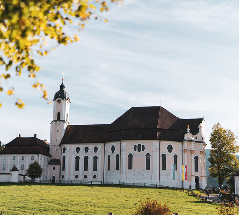 Das Bild zeigt eine friedliche Szene einer großen weißen Kirche mit einem rosa Dach, die majestätisch in der Mitte eines üppigen grünen Feldes steht. Die Kirche, mit ihrem hohen Turm und den vielen Fenstern, strahlt eine Atmosphäre der Ruhe und Ehrfurcht aus. Das Feld, lebendig und grün, erstreckt sich um die Kirche und schafft ein malerisches Setting. In der Ferne sind ein paar Bäume zu sehen, die zur natürlichen Schönheit der Szene beitragen. Die Kirche, mit ihrem auffälligen rosa Dach, hebt sich gegen das grüne Feld ab und zieht die Aufmerksam auf ihre architektonische Pracht. Die Gesamtansicht des Bildes suggeriert eine friedliche und idyllische Szene, vielleicht ein Ort der Anbetung und Reflexion.