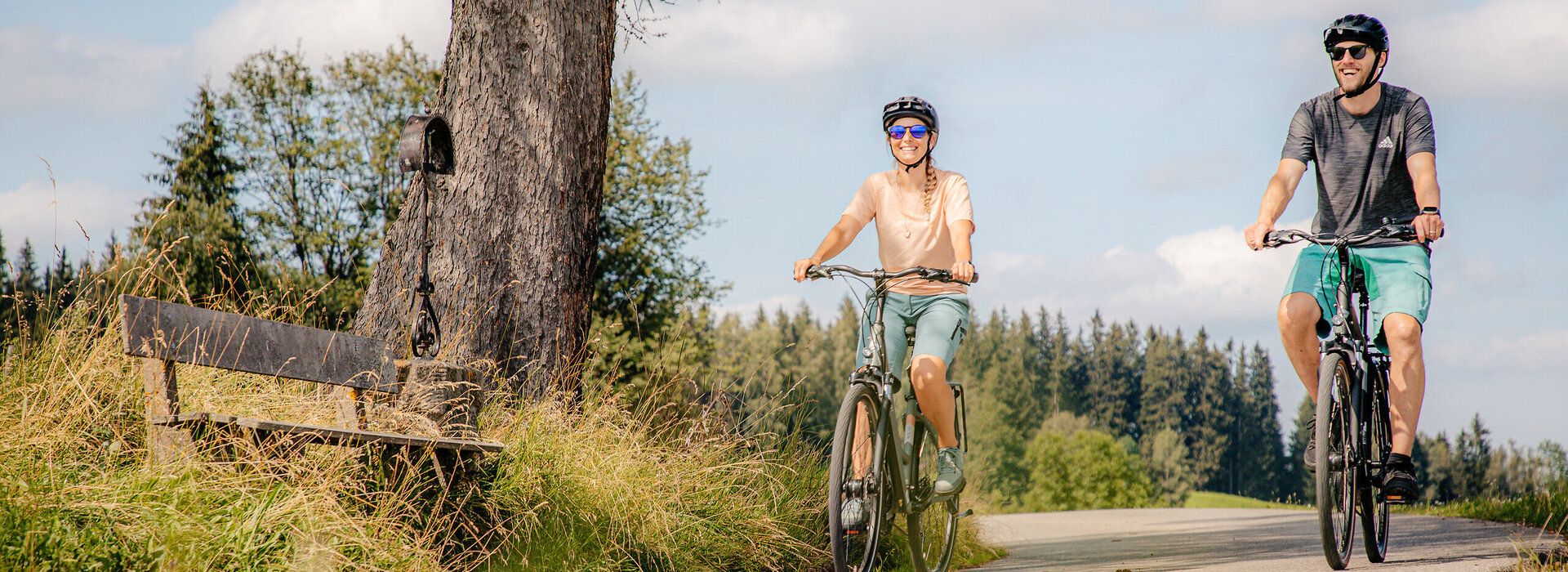 Das Bild zeigt einen Mann und eine Frau beim Fahrradfahren. Beide tragen Freizeitkleidung und einen Helm. Sie fahren auf den Betrachter zu. Beide lächeln und tragen Sonnenbrillen. Die Straße, auf der sie fahren ist asphaltiert. Wiesen und Wälder säumen die Straße. Am Straßenrand ist eine Wegkreuz aus Metall und eine Holzbank zu sehen.  Im Hintergrund erheben bewaldete Hügel. Der Himmel ist blau. Die Sonne scheint. 