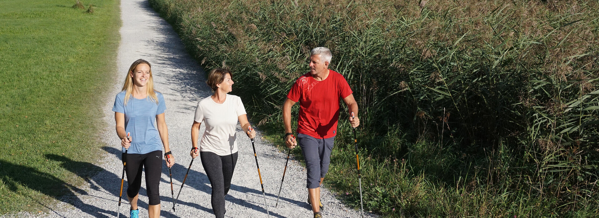 Das Bild zeigt drei Personen in Sportkleidung, die auf einem Uferweg laufen. Parallel zum Weg verläuft ein Schilfgürtel. Alle haben Nordic Walking Stöcke in den Händen. Sie lächeln. Im Hintergrund ist eine Ortschaft am Ufer des Sees zu sehen. Dahinter erheben sich hohe Berge und bewaldete Hügel.     