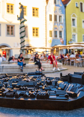 Die Abbildung zeigt einen belebten Stadtplatz mit Menschen. Vorne steht eine komplexe Metall-Konstruktion welche die Altstadt als Model wiedergibt. 