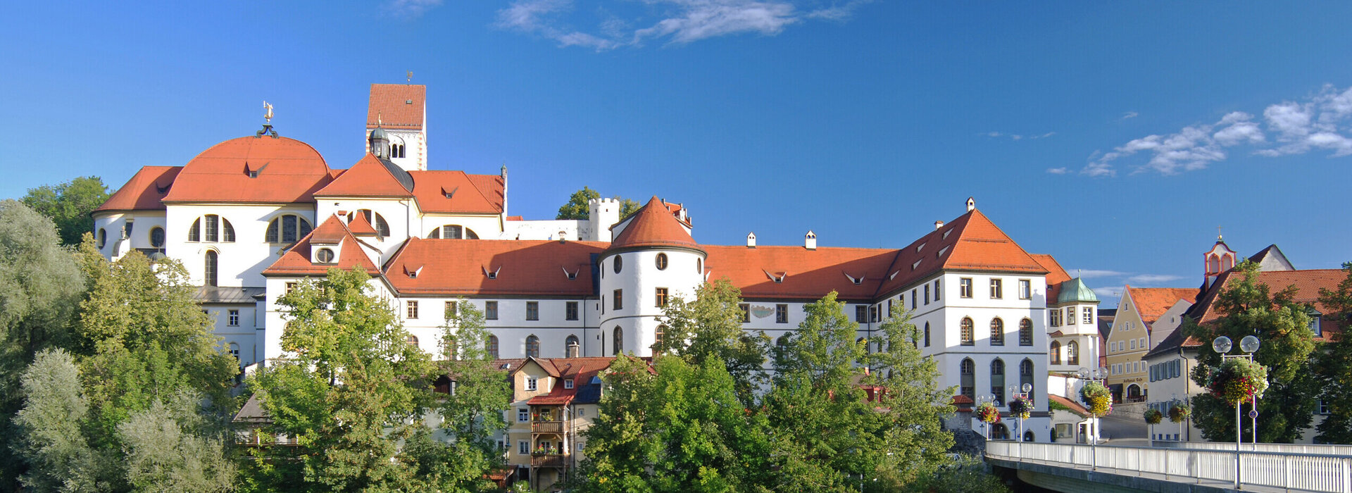 Das Bild zeigt ein großes, weißes Gebäude mit einem roten Dach, das in der Nähe eines Flusses, dem Lech steht. Das alte Kloster St. Mang ist von üppig grünen Bäumen umgeben, was eine friedliche und malerische Szene schafft.