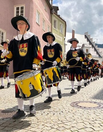 Das Bild zeigt einen Festzug. Eine Gruppe historisch gekleideter Personen mit gelb-schwarzen Trommeln laufen am Betrachter vorbei. Alle Personen haben Trommelstöcke in den Händen. Sie laufen durch eine Straße in Füssen. Im Hintergrund sind bunte Häuserfassaden zu erkennen. Viele Personen stehen am Straßenrand und beobachten den Festzug. Der Himmel ist blau mit weißen Wolken. Die Sonne scheint.