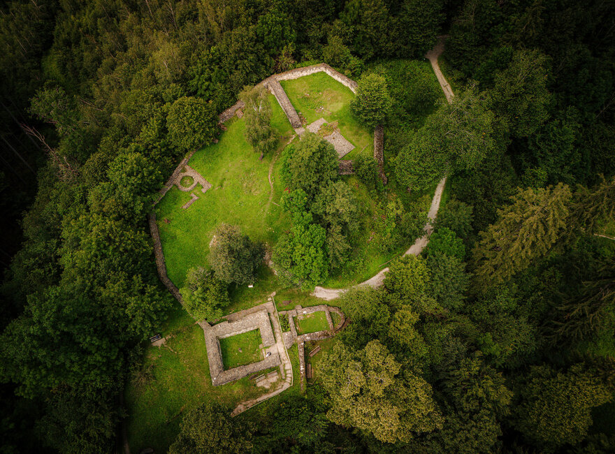 Das Bild zeigt die Mauerreste einer Burg. Das Foto ist aus der Vogelperspektive aufgenommen. Aus diesem Blickwinkel wirken die Mauerreste wie ein Grundriss, des ehemaligen Burggebäudes. Die Ruine steht auf einer Anhöhe und ist umgeben von dichtem Nadelwald. Es handelt sich um die Ruine der einstigen Burg Hopfen.