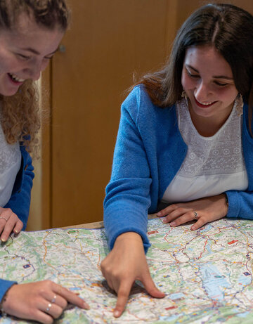 Das Foto zeigt drei Frauen, die sich über eine Karte beugen. Zwei Frauen zeigen etwas auf der Karte und lächeln. Zwei der drei Frauen tragen weiße Blusen und blaue Strickjacken.
