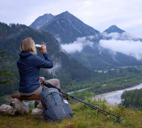 Das Bild zeigt eine Frau, die auf einem Holzscheit auf einer Anhöhe sitzt und ins Tal hinabblickt. Dort fließt ein Fluss. Sie trinkt aus einer Metallflasche und trägt Wanderkleidung. Neben ihr liegen Wanderstöcke. Im Hintergrund erheben sich bewaldete Berge. Der Himmel ist wolkenverhangen. Nebel zieht auf. 