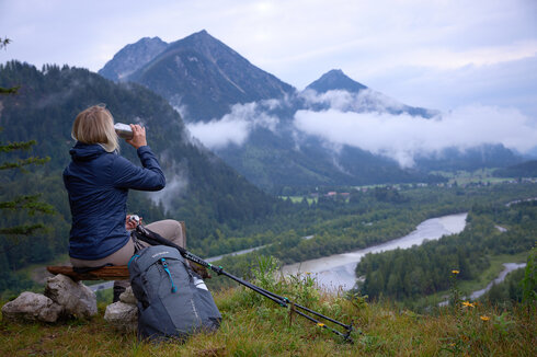 Das Bild zeigt eine Frau, die auf einem Holzscheit auf einer Anhöhe sitzt und ins Tal hinabblickt. Dort fließt ein Fluss. Sie trinkt aus einer Metallflasche und trägt Wanderkleidung. Neben ihr liegen Wanderstöcke. Im Hintergrund erheben sich bewaldete Berge. Der Himmel ist wolkenverhangen. Nebel zieht auf. 