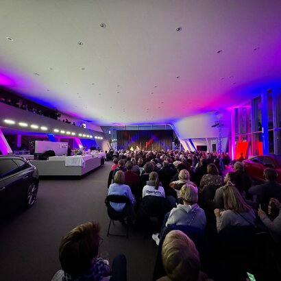 Das Bild zeigt mehrere Personen in Neoprenanzügen im knietiefen Wasser stehen. Alle tragen unterschiedliche Mützen und halten brennende Fackeln in den Händen. Es Dämmert. Es ist schon fast dunkel.  Auf der linken Seite des Bildes steht ein Auto. Auf einem Tresen sind Getränke und Gläser zu sehen. Im Hintergrund sieht man eine Bühne mit mehreren Musikern darauf. 