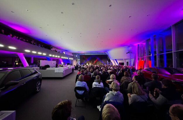Das Bild zeigt mehrere Personen in Neoprenanzügen im knietiefen Wasser stehen. Alle tragen unterschiedliche Mützen und halten brennende Fackeln in den Händen. Es Dämmert. Es ist schon fast dunkel.  Auf der linken Seite des Bildes steht ein Auto. Auf einem Tresen sind Getränke und Gläser zu sehen. Im Hintergrund sieht man eine Bühne mit mehreren Musikern darauf. 