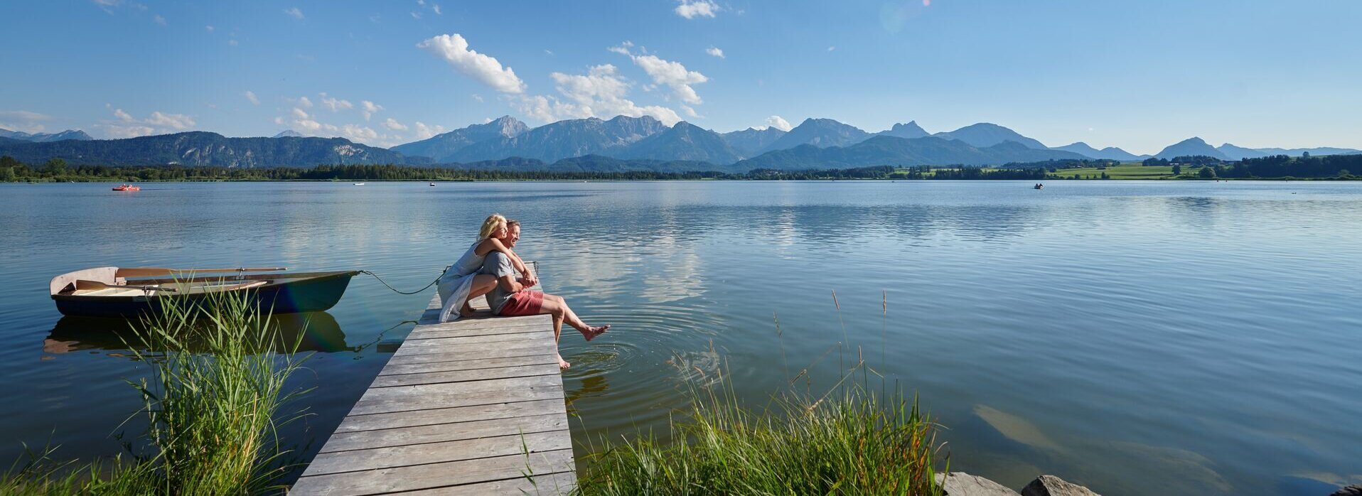 Auf dem Bild ist eine idyllische Szene am Hopfensee zu sehen. Es ist Sommer. Eine Frau kniet hinter einem Mann auf einem Holzsteg und umarmt ihn von hinten. Er sitzt auf dem Holzsteg. Seine Beine baumeln über dem Rand des Stegs herunter. Seine Füße berühren leicht die Wasseroberfläche des tiefblauen Sees. Beide blicken in die Ferne und lächeln. Der Holzsteg ragt in das ruhige Wasser des Hopfensees hinein, welcher von Bergen umgeben ist. Der Himmel ist blau und nur leicht durchzogen von wenigen Wolken. In der Ferne sind ein paar Ruder- und Tretboote auf dem Wasser zu sehen.