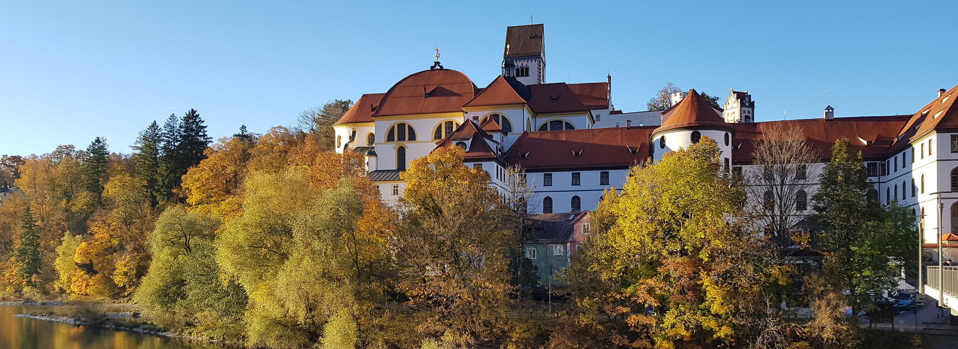 Das Bild zeigt den Blick auf das ehemalige Kloster St. Mang in Füssen im Allgäu vom gegenüberliegenden Lechufer aus. Der brocke Klosterbau erhebt sich über dem herbstlich gefärbten Baumbestand entlang des Flusses Lech. 