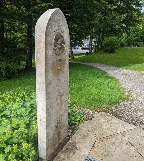  Das Bild zeigt ein schmales, längliches, steinernes Gebilde, das wie ein Denkmal aussieht. Die Struktur ist rechteckig geformt, mit einer gewölbten Oberseite. Es handelt sich um den Arnika Trinkbrunnen. Dieser befindet sich in einem parkähnlichen Areal, umgeben von üppigem grünem Gras und Bäumen. Der Brunnen ist mit einer Metallplatte verziert, die im oberen Drittel befestigt ist. Aus der Metallplatte ragt ein Wasserhahn. In die Metallplatte ist die Struktur einer Arnika-Blüte eingarbeitet.  Die Platte trägt die Inschrift "Arnika". Die gesamte Szene suggeriert eine friedliche und ruhige Umgebung. 
