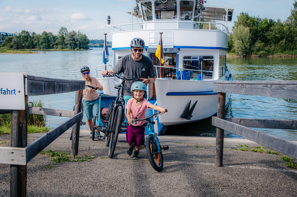 Das Bild fängt eine ruhige Szene an einer Anlegestelle ein. Eine Familie von drei, bestehend aus einem Mann, einer Frau und einem Kind, steht auf der Anlegestelle, wobei der Mann und das Kind auf Fahrrädern stehen. Der Mann hält das Fahrrad des Kindes, während das Kind das der Frau hält. Sie tragen alle Helme, was auf Sicherheitsvorkehrungen hindeutet.  Im Hintergrund liegt ein großes weißes Boot vertäut. Das Boot ist mit einer blauen und gelben Flagge verziert, was dem Bild einen Farbtupfer verleiht. Die Anlegestelle ist von üppig grünen Bäumen umgeben, was eine malerische Kulisse schafft. Die Familie scheint ihre Zeit zusammen zu genießen, vielleicht bereiten sie sich auf eine Fahrradtour vor oder sie nehmen einfach die schöne Umgebung wahr.