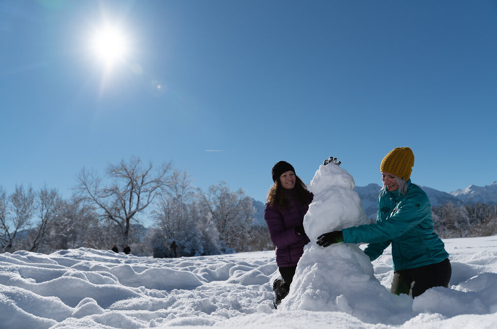  Das Bild fängt einen fröhlichen Moment in einer schneebedeckten Landschaft ein. Zwei Frauen stehen im Schnee, ihre Körper sind teilweise darin versunken. Sie sind mit der fröhlichen Aktivität des Schneemannbaus beschäftigt. Die Frau auf der linken Seite hält eine große Schneeball, die Frau auf der rechten Seite eine kleinere. Sie beide tragen winterliche Kleidung, wobei die Frau auf der rechten Seite ein lebhaft blaues Jacket trägt. Der Schneemann, den sie bauen, befindet sich gerade im Aufbau-Prozess, wobei nur die Basis sichtbar ist. Die Kulisse des Bildes ist eine atemberaubende Aussicht einer schneebedeckten Bergkette unter einem klaren blauen Himmel. Die Sonne scheint hell, und wirft ein warmes Licht auf die Szene. Das Bild vermittelt ein Gefühl von Spaß, Kameradschaft und der einfachen Freude, Zeit im Winterdraußen zu verbringen.