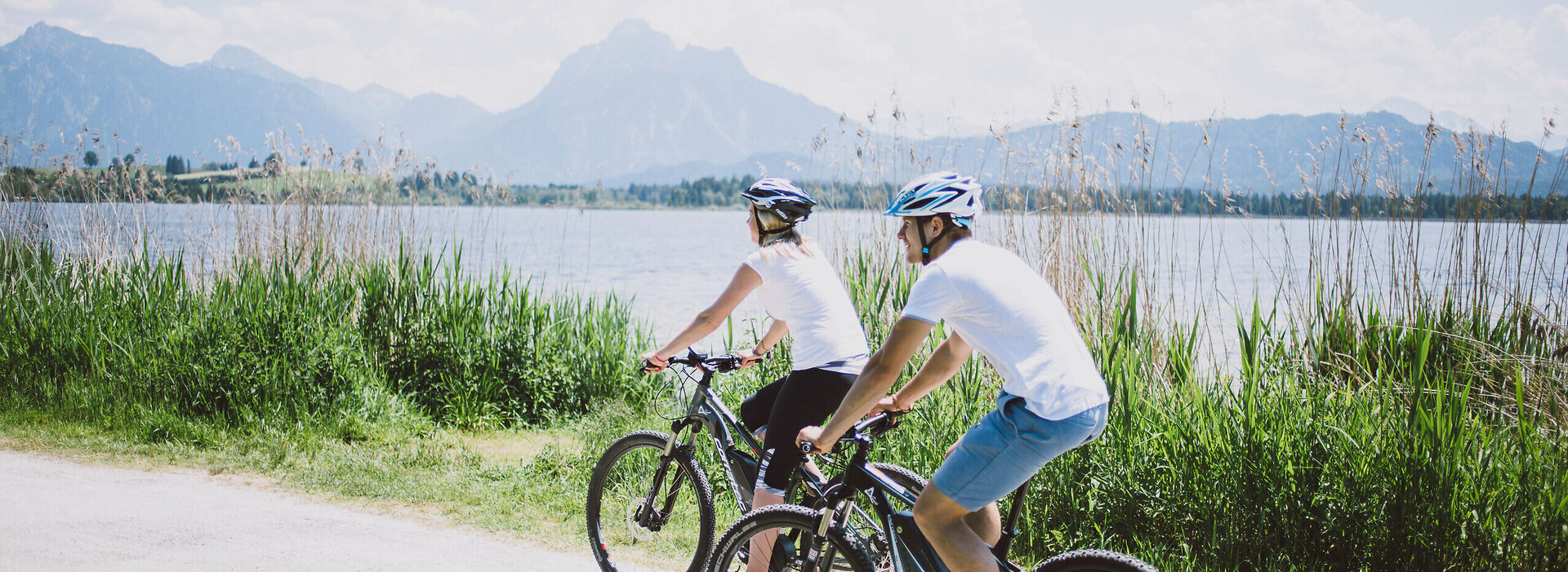 Das Bild zeigt einen Mann und eine Frau, die Fahrrad auf einem Fahrradweg entlang des Hopfensees fahren. Der Fahrradweg ist führt an grünen Wiesen entlang. Im Hintergrund sieht man den Hopfensee und die Berge. Die Radfahrer sind in lässiger Kleidung und mit Helm gekleidet. Beide tragen ein weißes T-Shirt und eine kurze Hose. Das Bild vermittelt eine entspannte Fahrradfahrt.