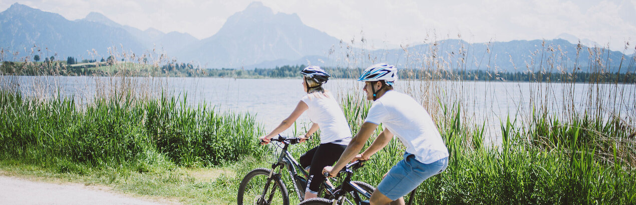 Das Bild zeigt einen Mann und eine Frau, die Fahrrad auf einem Fahrradweg entlang des Hopfensees fahren. Der Fahrradweg ist führt an grünen Wiesen entlang. Im Hintergrund sieht man den Hopfensee und die Berge. Die Radfahrer sind in lässiger Kleidung und mit Helm gekleidet. Beide tragen ein weißes T-Shirt und eine kurze Hose. Das Bild vermittelt eine entspannte Fahrradfahrt.