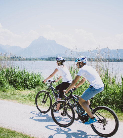 Das Bild zeigt einen Mann und eine Frau, die Fahrrad auf einem Fahrradweg entlang des Hopfensees fahren. Der Fahrradweg ist führt an grünen Wiesen entlang. Im Hintergrund sieht man den Hopfensee und die Berge. Die Radfahrer sind in lässiger Kleidung und mit Helm gekleidet. Beide tragen ein weißes T-Shirt und eine kurze Hose. Das Bild vermittelt eine entspannte Fahrradfahrt.