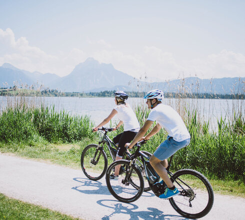 Das Bild zeigt einen Mann und eine Frau, die Fahrrad auf einem Fahrradweg entlang des Hopfensees fahren. Der Fahrradweg ist führt an grünen Wiesen entlang. Im Hintergrund sieht man den Hopfensee und die Berge. Die Radfahrer sind in lässiger Kleidung und mit Helm gekleidet. Beide tragen ein weißes T-Shirt und eine kurze Hose. Das Bild vermittelt eine entspannte Fahrradfahrt.