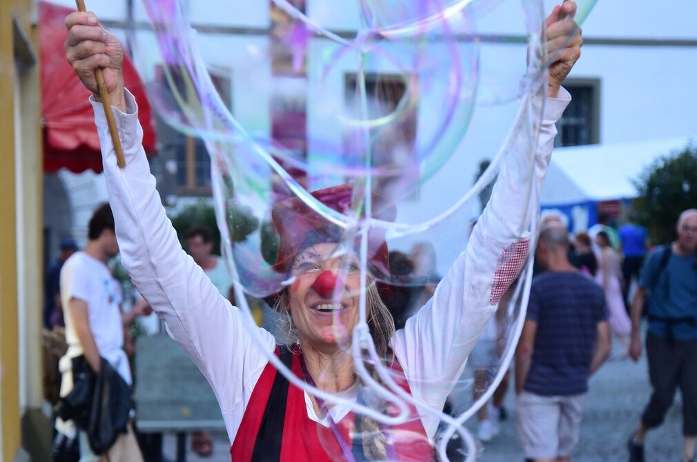 Das Bild zeigt eine Straßenkünstlerin. Sie ist als Clown verkleidet und geschminkt und trägt eine rote Nase. In ihren Händen hält sie zwei Holzstäbe in die Höhe. Vor ihr schweben riesige Seifenblase. Im Vordergrund recken Kinder ihre Hände und Arme in die Höhe. Die Sonne scheint. 
