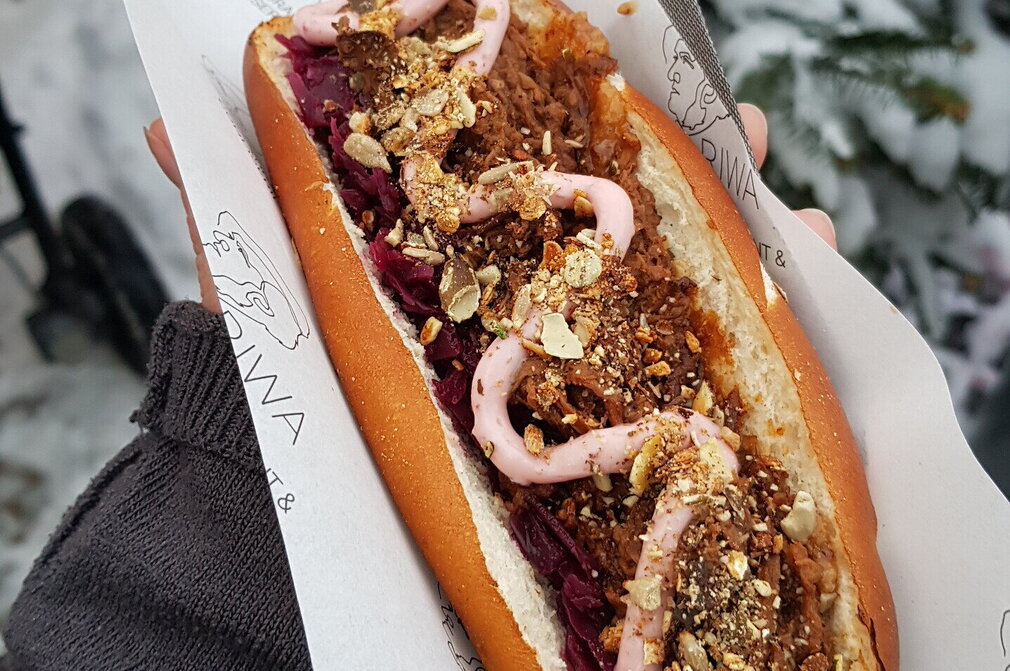 Das Bild zeigt eine längliche Semmel. Sie ist der Länge nach aufgeschnitten und enthält eine rot- bräunliche Masse. Garniert mit Nüssen und einer rosa Soße, die in Schlangenlinien auf der Masse angebracht wurde. Die Semmel liegt auf einer Serviette. Eine Hand hält die Semmel. 