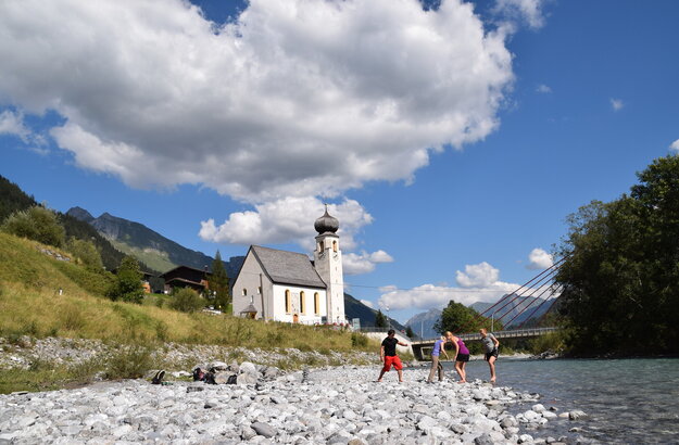 Das Bild zeigt vier Personen, die in einem Bachbett stehen. Das Ufer es Bachbetts ist mit groben Steinen und Kies bedeckt. Sie haben die Schuhe ausgezogen und treten ins Wassern. Im Hintergrund steht eine Kirche mit Kirchturm. Dahinter erheben sich die Berge. Der Himmel ist blau. Die Sonne scheint. 