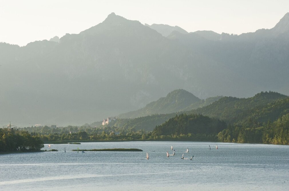 Das Bild zeigt den Weißensee, eingebettet in eine bergige Landschaft. Der See ist von einem tiefen Blau und zeigt mehrere Windsurfer, deren weiße Segel das Licht einfangen. Die Surfer sind über das Wasser verstreut, manche näher am Vordergrund, während andere weiter entfernt sind. Die Berge erheben sich majestätisch im Hintergrund, wobei ihre Gipfel den Himmel berühren. Der Himmel selbst ist hell und blau.