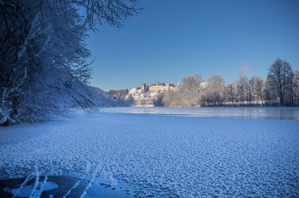 Das Bild zeigt ruhige Winterlandschaft. Im Vordergrund dominiert der teilweise zugefrorene Fluss Lech. Der zugefrorene Teil ist mit weißen Eisblumen und Eiskristallen bedeckt. Der Fluss nimmt den größten Teil des Bildes ein, seine Wasseroberfläche spiegelt das blasse Blau des Himmels. In der Ferne thront majestätisch das Hohe Schloss und das ehemalige Kloster St. Mang auf einer Anhöhe. Auch die Bäume, die am Ufer stehen, sind mit Schnee bedeckt. 