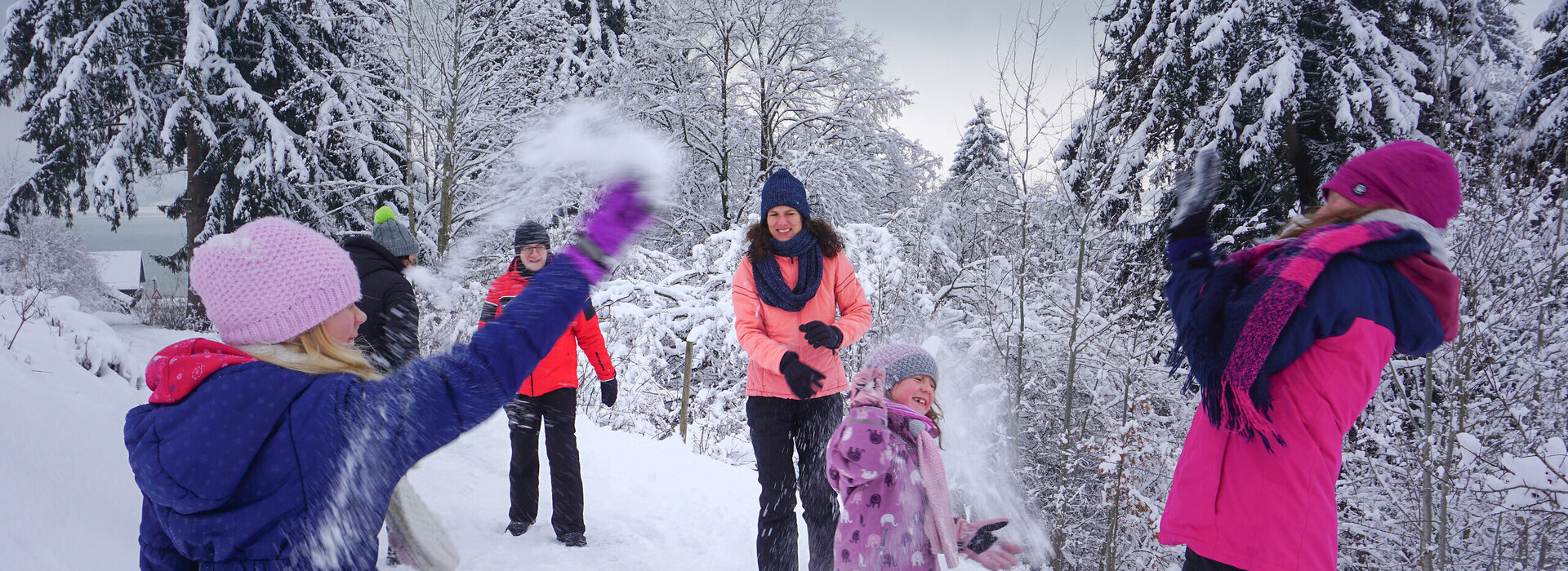 Das Bild fängt eine fröhliche Winterszene in einem schneebedeckten Wald ein. Eine Gruppe von Menschen, die in bunten Winterbekleidungen gekleidet sind, genießt den Schnee. Einige von ihnen werfen Schneebälle, was eine spielerische Atmosphäre schafft. Die Bäume im Hintergrund sind mit Schnee bedeckt, was zur winterlichen Atmosphäre beiträgt. Der Himmel über ihnen ist wolkenverhangen, was auf einen kalten und verschneiten Tag hinweist. Die Menschen stehen auf einem schneebedeckten Weg, der durch den Wald führt. Die gesamte Szene vermittelt ein Gefühl von Spaß und Kameradschaft, da die Gruppe von Freunden oder Familienmitgliedern Zeit miteinander in den Schnee verbringt.