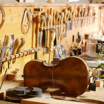Man blickt auf eine hölzerne Werkbank, an deren Hinterwand unzählige Feilen, Schnitzmesser und andere Werkzeuge befestigt sind. Auf der Werkbank liegt der Korpus einer Geige, neben weiteren Utensilien für den Geigenbau. Der Raum ist hell und einladend.  