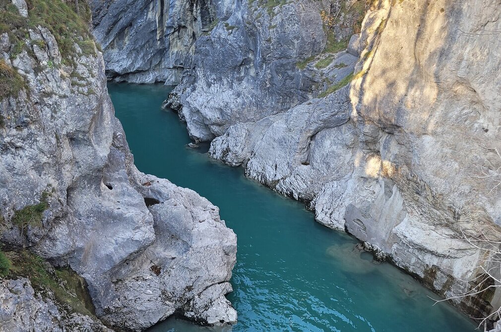 Das Bild zeigt einen atemberaubenden Blick auf einen Fluss, der durch eine felsige Schlucht fließt. Der Fluss Lech, mit seinem lebendigen blau-grünen Wasser, steht im Mittelpunkt des Bildes. Er schlängelt sich durch die Schlucht, eingebettet zwischen hochragenden Felsformationen auf beiden Seiten. Die Felsen, die in verschiedenen Grau- und Weißtönen variieren, geben der Szene ein Gefühl der Wildheit. Das Licht der Sonne bricht durch die Schlucht und wirft ein warmes Glitzern auf die Felsen und den Fluss. 
