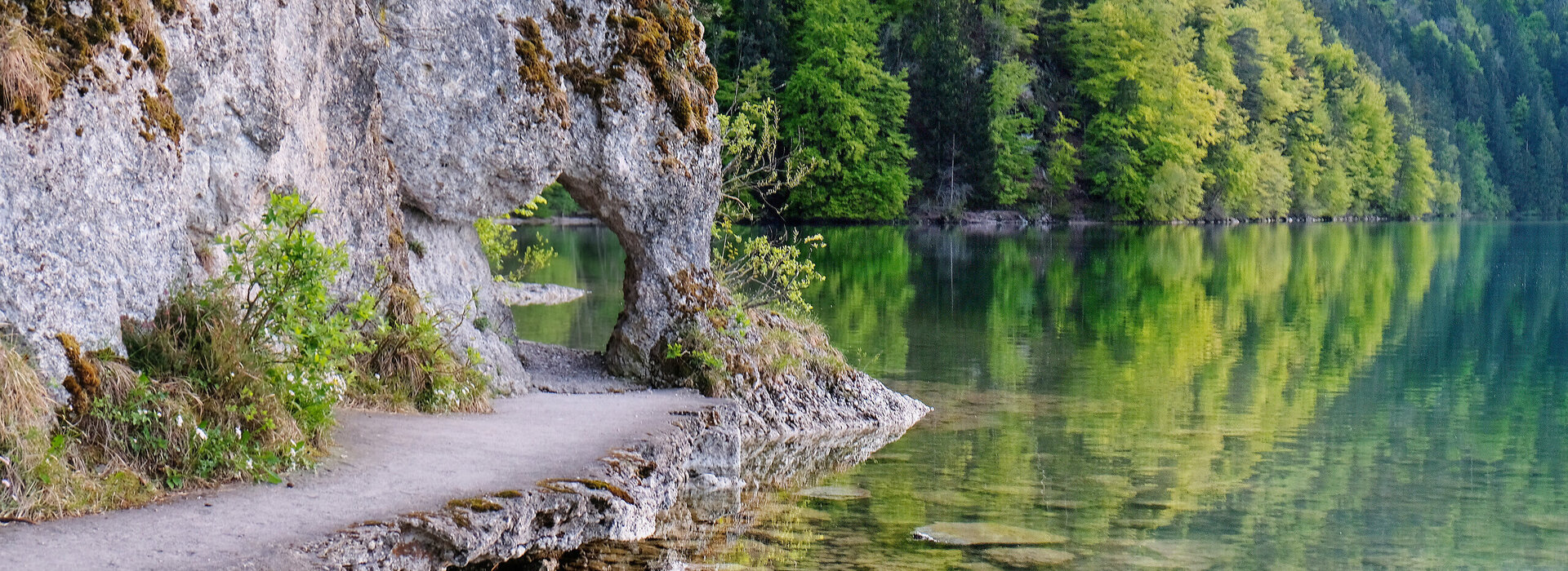 Das Bild zeigt einen See. Im linken Bildrand ist der Uferbereich zu sehen, an dem eine Felswand emporsteigt. Der Felsen bildet ein Art „Tor“, durch das sich der Weg fortsetzt. Der Uferweg vor dem „Felsentor“ ist befestigt. Das Wasser ist klar. Im Hintergrund ragt der Bergwald in die Höhe. Die Bäume sind in unterschiedliche Grüntöne getaucht. Im Vordergrund des Bildes kann man den steinigen Seeboden durch das klare Wasser sehen.   