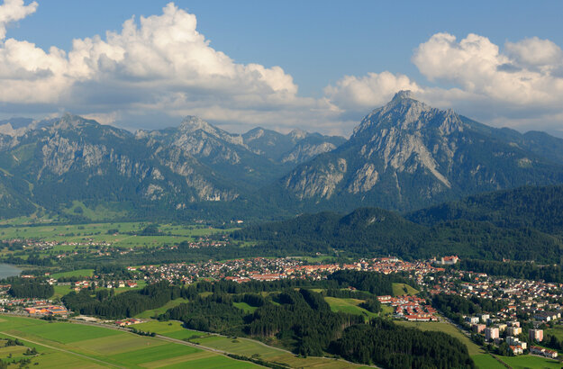 Das Foto zeigt Füssen von oben, umrahmt von Bergen, Hügeln, grünen Wiesen und Wäldern. Auf einer Anhöhe über Füssen thront das Hohe Schloss. Im Hintergrund ist das Schloss Neuschwanstein zu sehen, dass auf einer Erhebung vor den Bergen steht. 