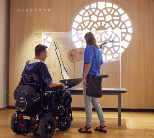 Das Bild zeigt einen Mann und eine Frau in einer Museumssituation. Die Frau, die steht, zeigt auf eine Ausstellung, während der Mann, der im Rollstuhl sitzt, auf sie schaut. Beide Personen tragen blaue Hemden. Das Museum verfügt über eine Vielzahl von Ausstellungen, darunter eine Darstellung von Musikinstrumenten und eine von Büchern. Das gesamte Ambiente des Museums ist ruhig und bildend.