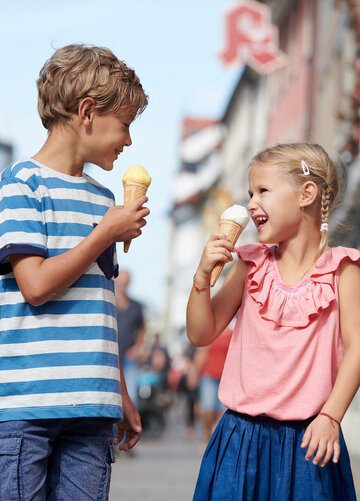 Das Bild fängt eine herzliche Szene von zwei Kindern ein, einem Jungen und einem Mädchen, die einen sonnigen Tag in einer europäischen Stadt genießen. Der Junge trägt ein gestreiftes Hemd in Blau und Weiß und hält einen gelben Eis in der Hand. Das Mädchen, das ein pinkfarbenes Oberteil trägt, hält ein weißes Eis. Sie beide lächeln, ihre Gesichter strahlen vor Freude. Die Stadt um sie herum ist lebendig und geschäftig. Im Hintergrund sind Gebäude, Bäume und Menschen zu sehen, die ihren Alltag verbringen. Der Himmel über ihnen ist klar und blau, was zur allgemeinen Lebensfreude der Szene beiträgt. Die Freude der Kinder ist greifbar, was das Bild zu einer wirklich herzerwärmenden Darstellung macht.