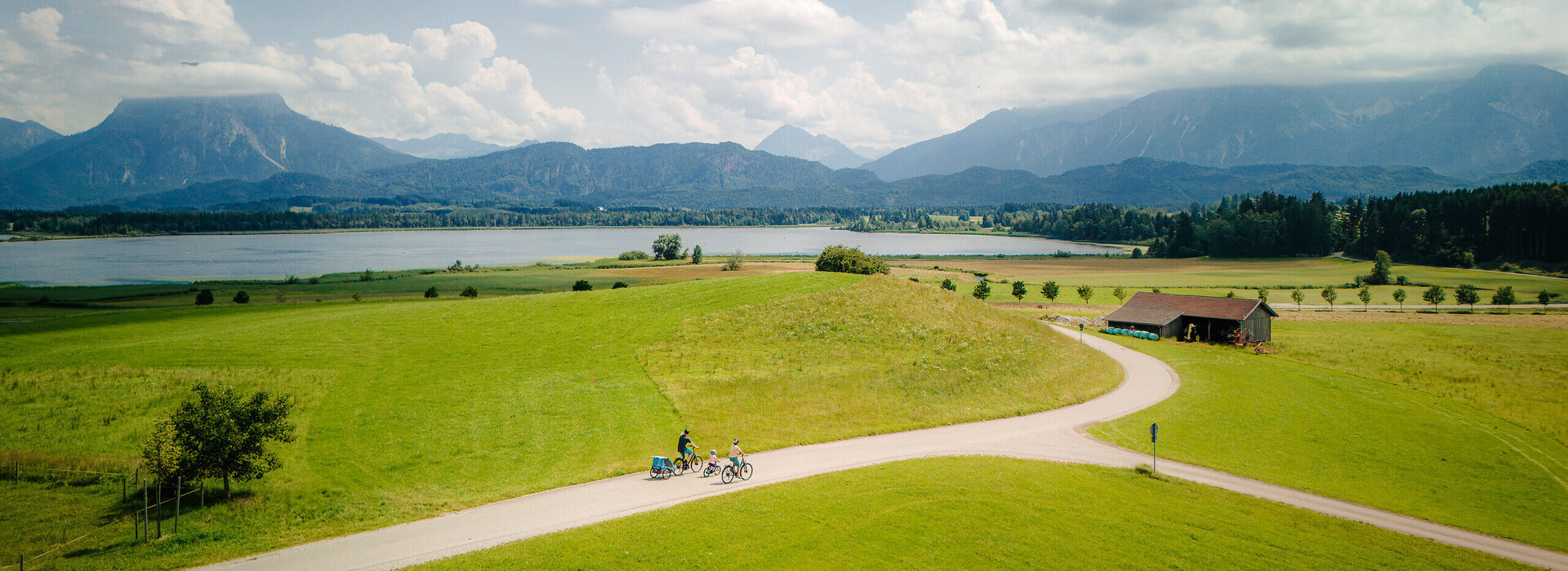 Das Bild zeigt im Vordergrund eine asphaltierte Straße, die durch eine Wiese führt. Auf der Wiese fahren zwei Erwachsene und ein Kind Fahrrad. Am Fahrrad einer Person ist ein Fahrradanhänger befestigt. Die Straße scheint zum dahinterliegenden See zu führen. Im Hintergrund erheben sich Berge und grün, bewaldete Hügel. Der Himmel ist blau. Die Sonne scheint. 