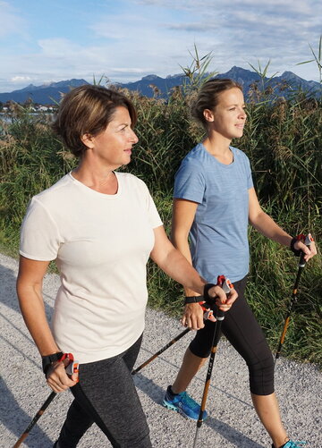 Das Bild zeigt zwei Frauen in Sportkleidung, die auf einem Uferweg laufen. Parallel zum Weg verläuft ein Schilfgürtel. Beide haben Nordic Walking Stöcke in den Händen. Sie lächeln. Im Hintergrund ist eine Ortschaft am Ufer des Sees zu sehen. Dahinter erheben sich hohe Berge und bewaldete Hügel.     