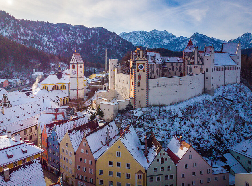 Das Bild fängt die atemberaubende Sicht auf das mittelalterliche Städtchen Füssen in Deutschland ein. Die Stadt liegt auf einem Hügel, von majestätischen Bergen umgeben. Die Architektur der Stadt ist durch bunte Gebäude geprägt, jedes mit einem einzigartigen Charme. Die Gebäude sind mit Schnee verziert, was eine Prise Winter in die Szene bringt